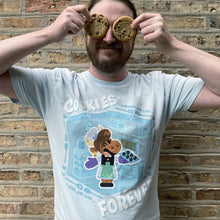 Person wearing a t-shirt with a cartoon character and text, holding cookies up to their eyes against a brick wall.